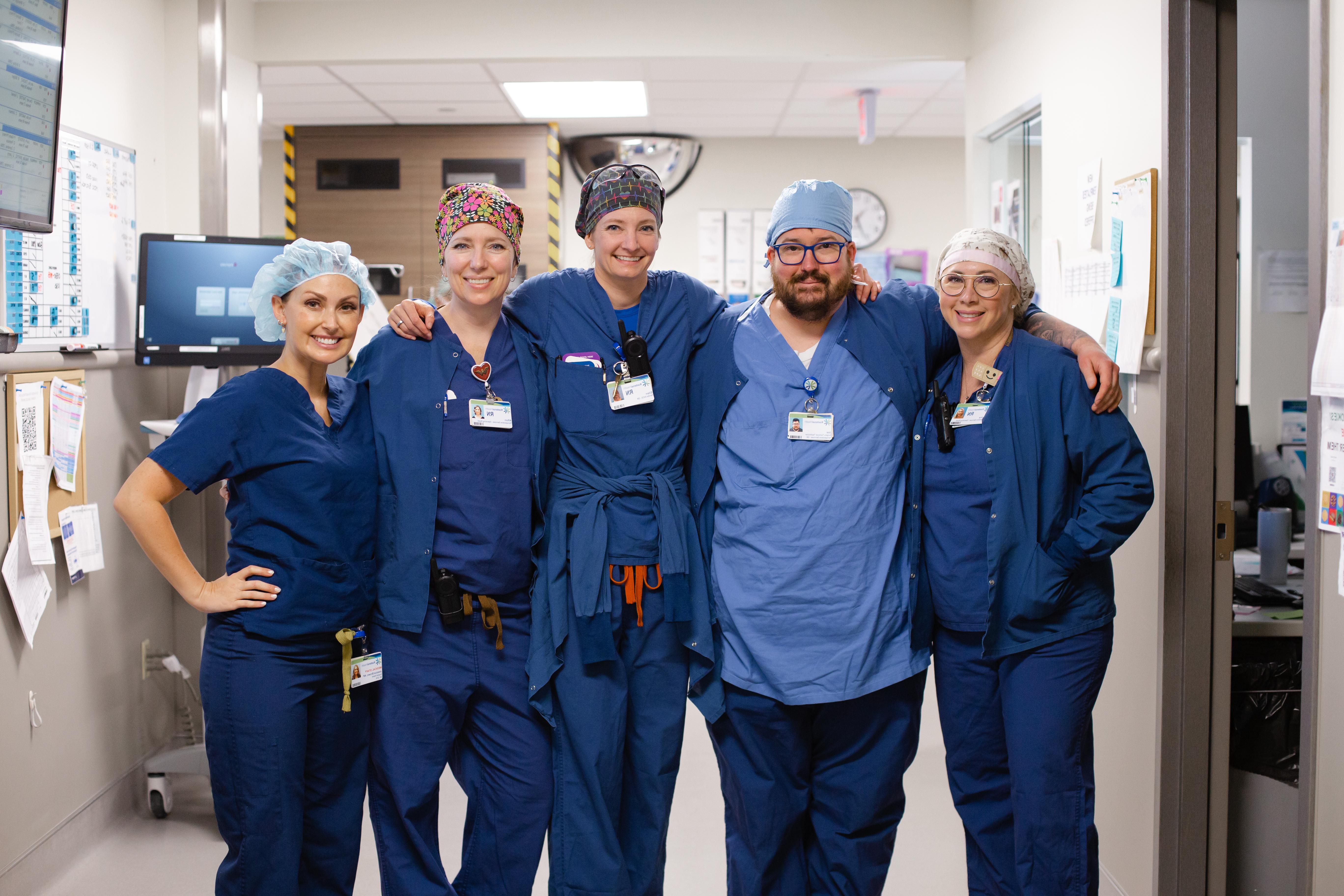 5 nurses in blue scrubs stand together with arms around each other
